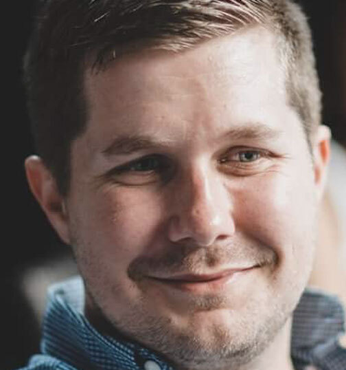 Britt Clark, a white man with short dirty blonde or brown hair, looking away from the camera and wearing a blue-checkered button up.