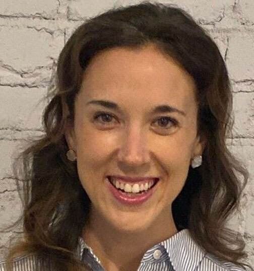 Emily Ellis, a white woman with shoulder-length, wavy brown hair, wearing a button-up with thin vertical stripes and smiling.