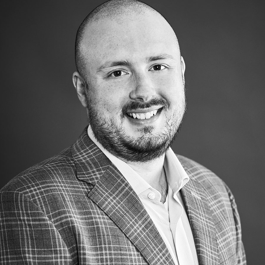 In black and white, Kalın Kingsland, a bald white man, wearing a plaid suit jacket and smiling at the camera.